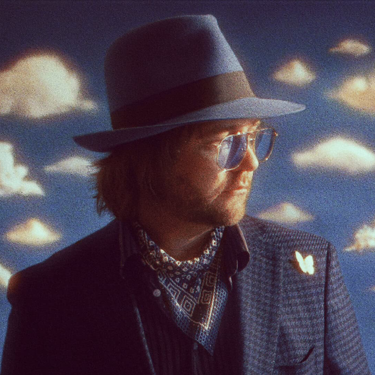 male musician with tinted glasses and hat with cloud background