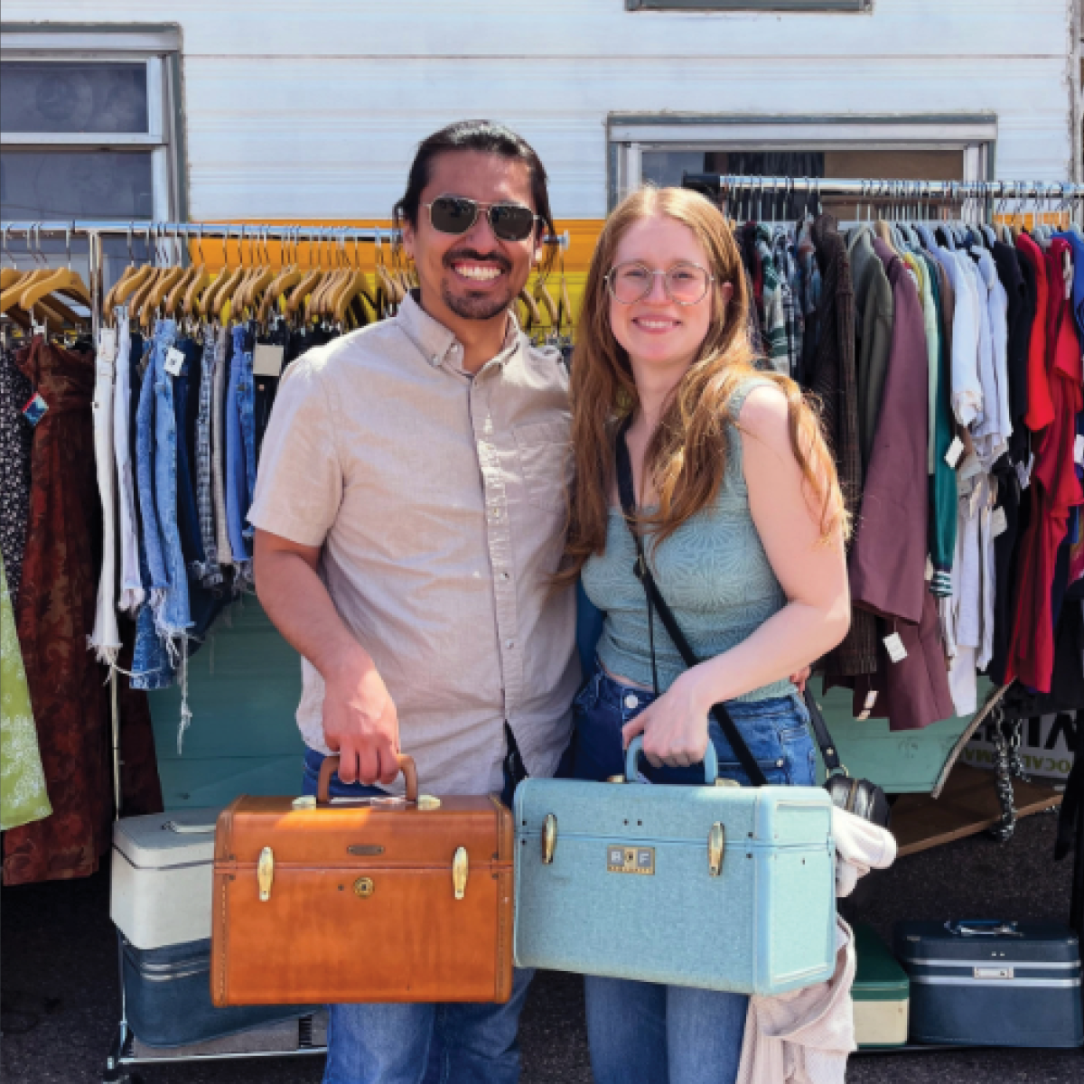 a young hip couple holding 2 small vintage suitcases with a rack of vintage clothes in the background