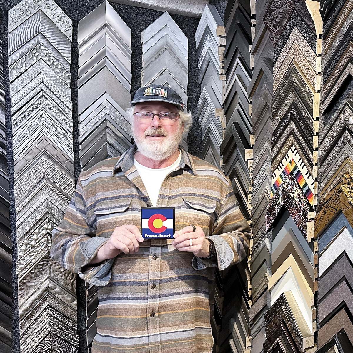 owner of frame shop standing in front of a wall of frames holding a small colorado flag