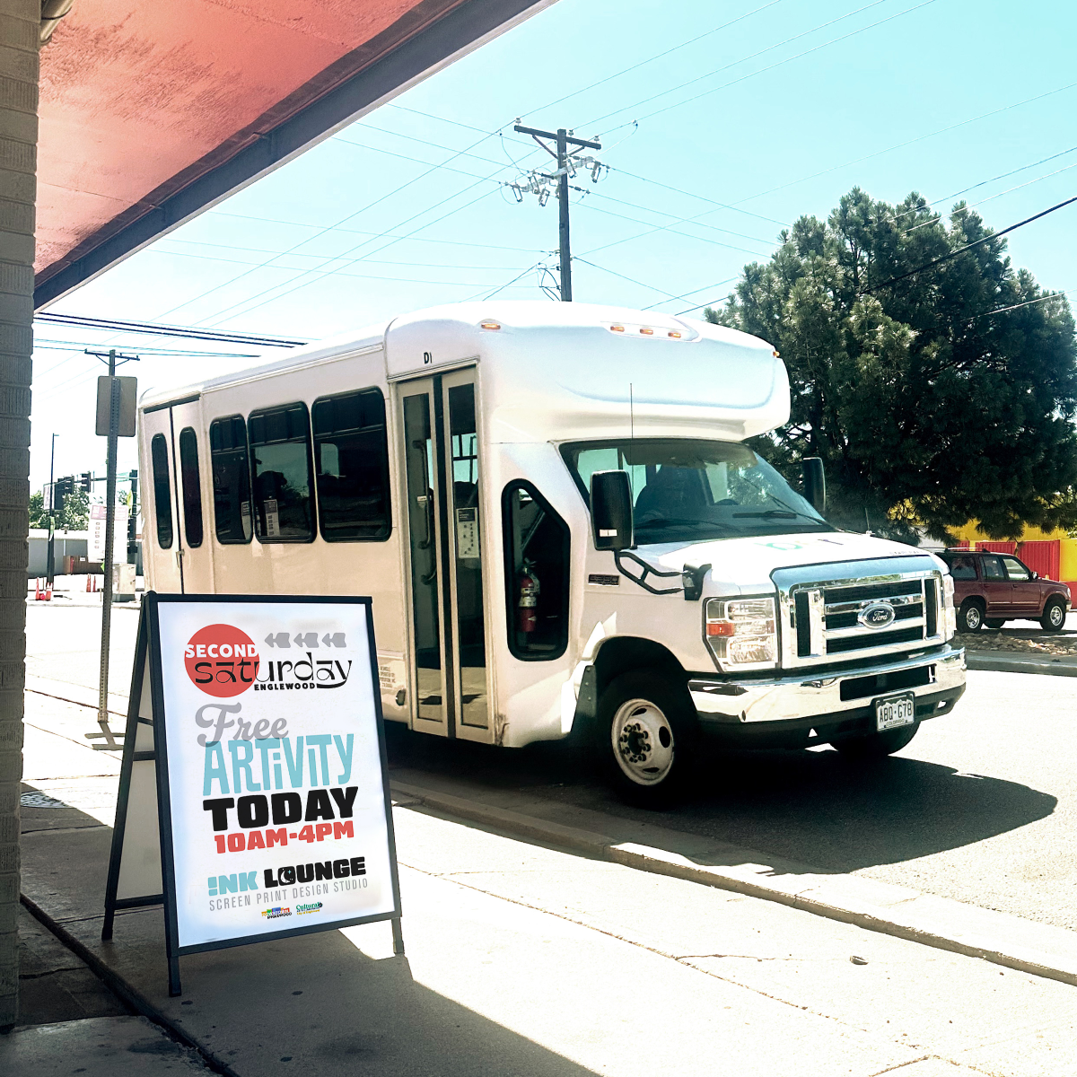 a white shuttle bus with an A-frame sign on the sidewalk promoting 2nd Saturdays